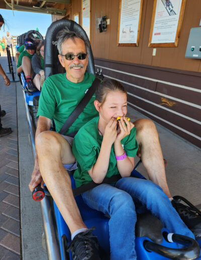 A smiling man and a giggling girl, both in green shirts, are seated in a blue amusement park ride car.