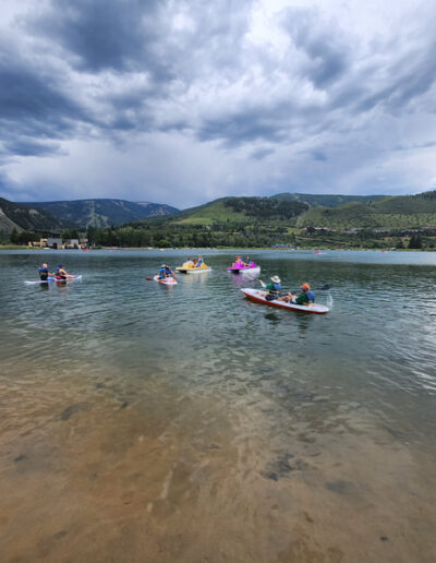 Campers and guides on paddle boards and paddle boats on a calm water under a dramatic cloudy sky, surrounded by green, hilly landscape.