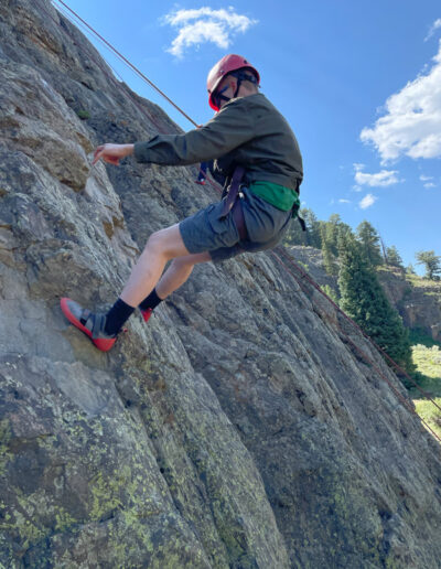 Camper rock climbing a steep, rugged rock in a harness and red helmet.
