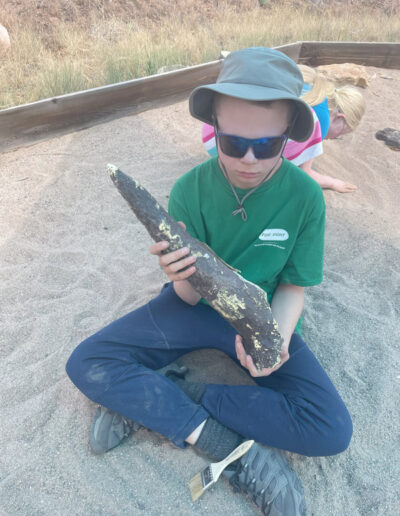 Boy with sunglasses and green shirt sitting on sand holding a large pointed rock or piece of wood.