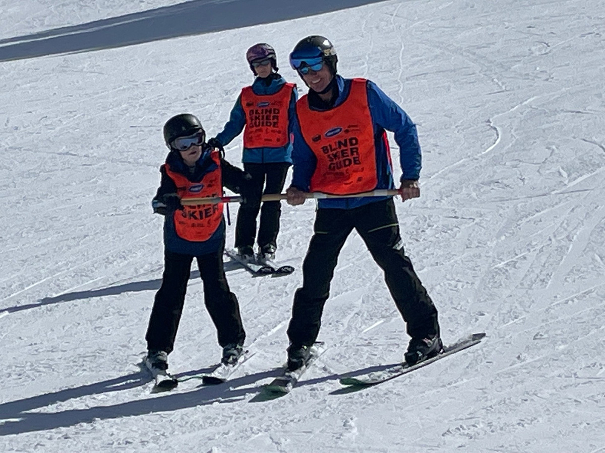 Guide working with blind child using a bamboo pole to get down the ski run